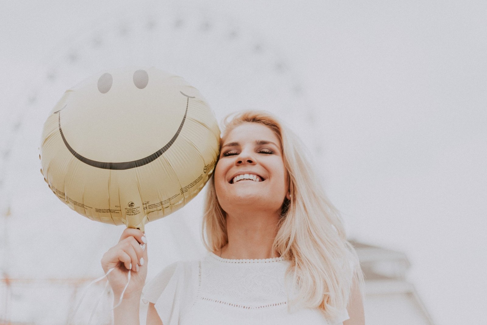 Woman holding a smiling face balloon, she is also smiling and there is a Ferris wheel in the background.